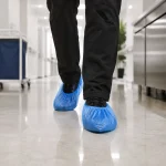 Person wearing blue disposable shoe covers walking down a clean, bright hallway (likely a hospital/ lab environment).