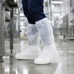 Person wearing disposable boot covers over blue pants in a clean industrial facility to prevent contamination or dirt on floors.