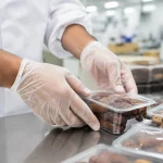 Close-up of gloved hands sealing a clear plastic container of dates on a stainless steel worktable in a clean food-processing area.
